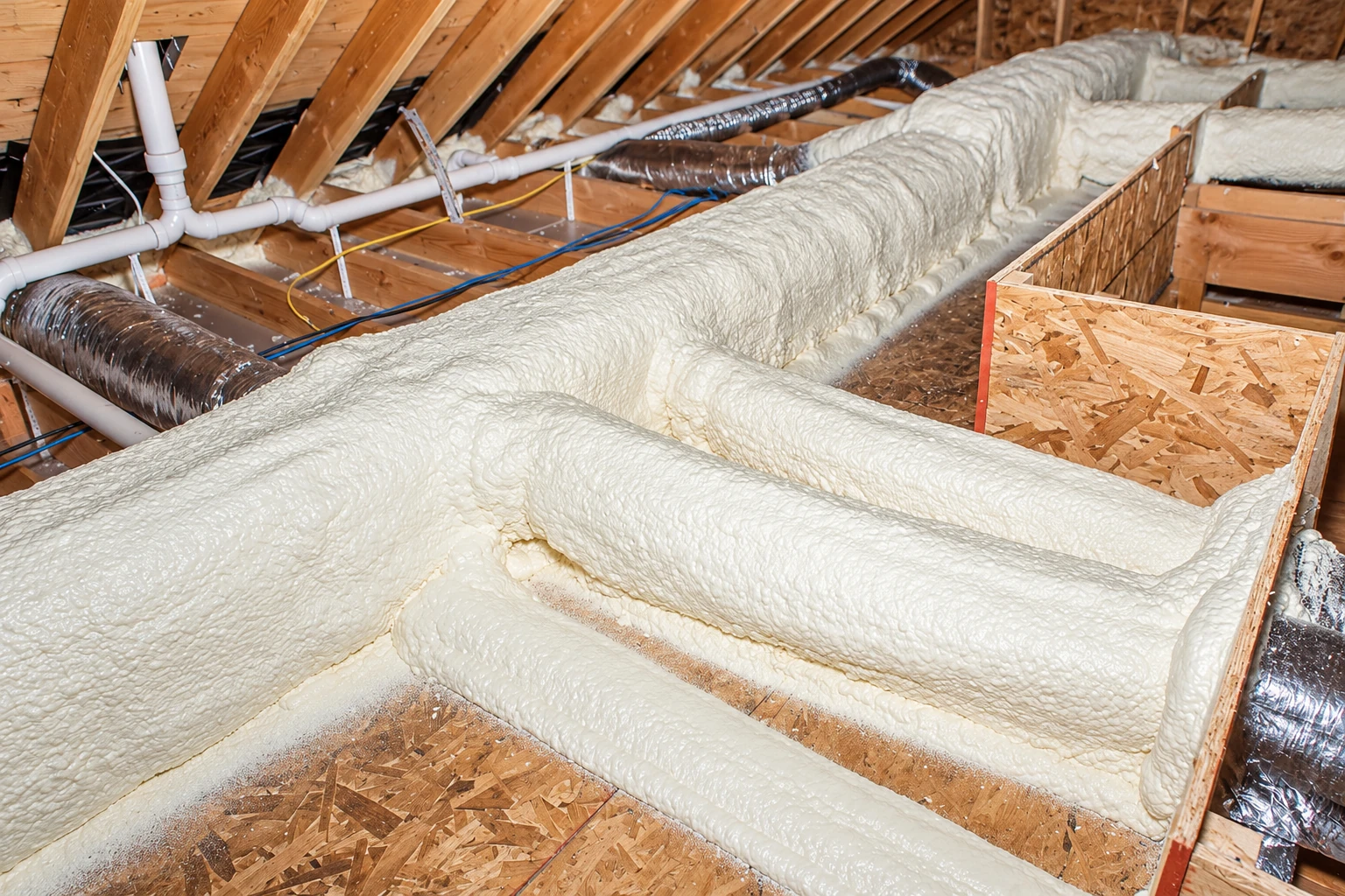 Attic framing coated in thick white foam around ducts, pipes, and wiring.