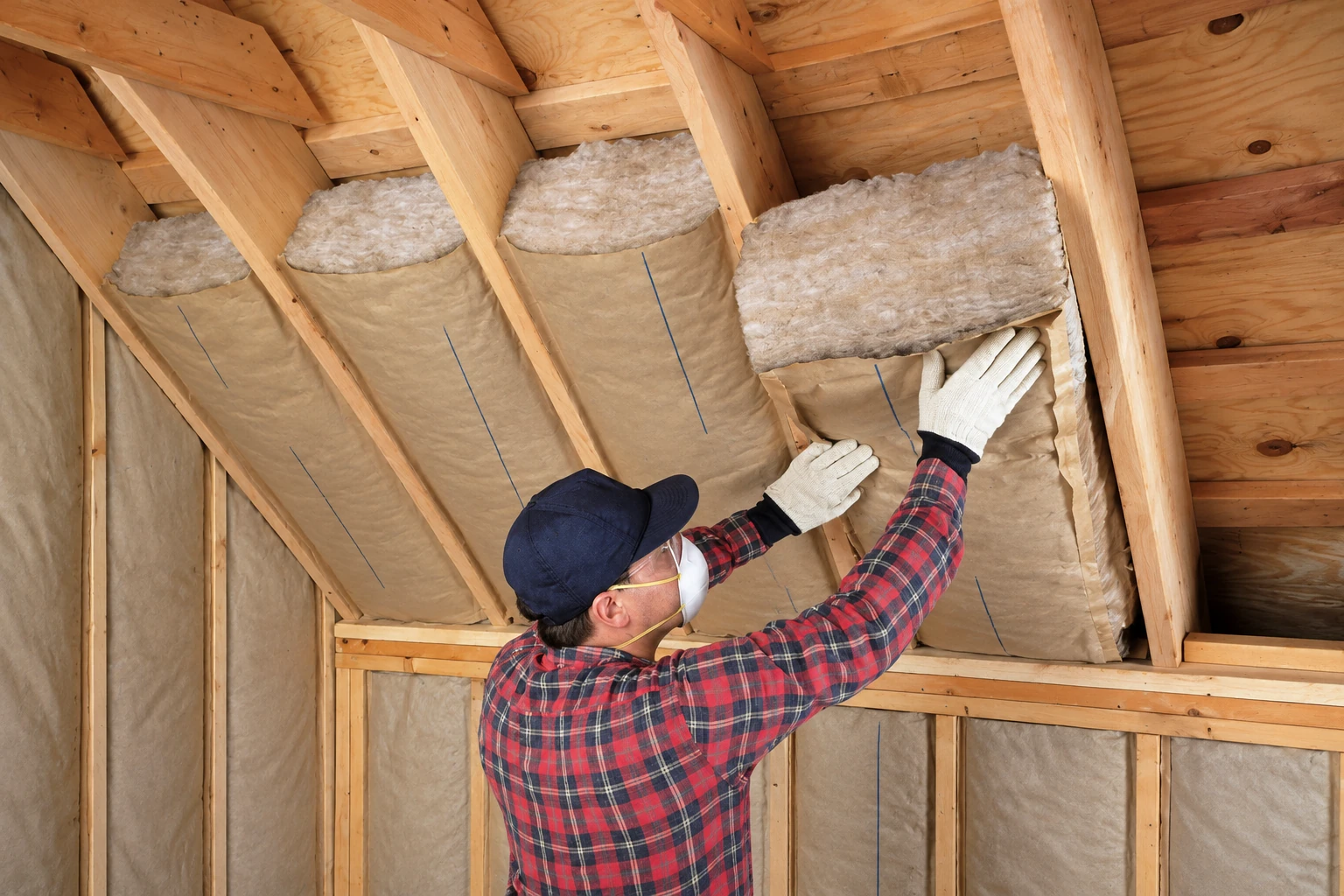 A worker in gloves and a respirator fits fiberglass batts between attic rafters.
