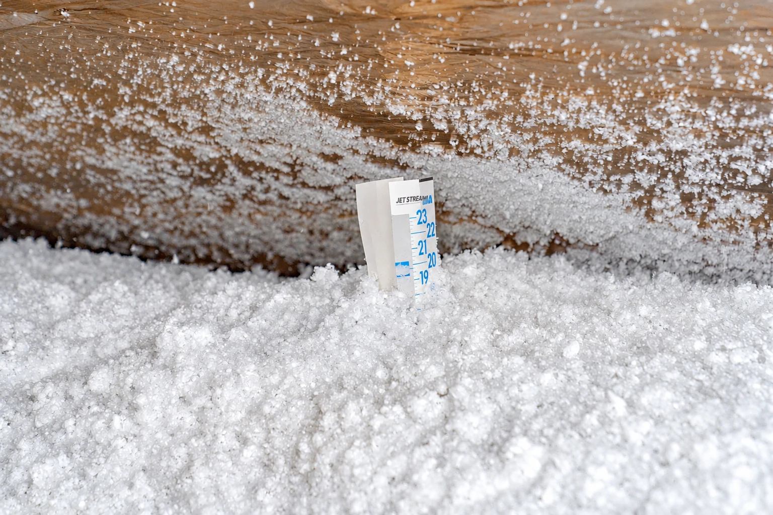 White blown-in insulation covers an attic floor around a depth-measuring marker.