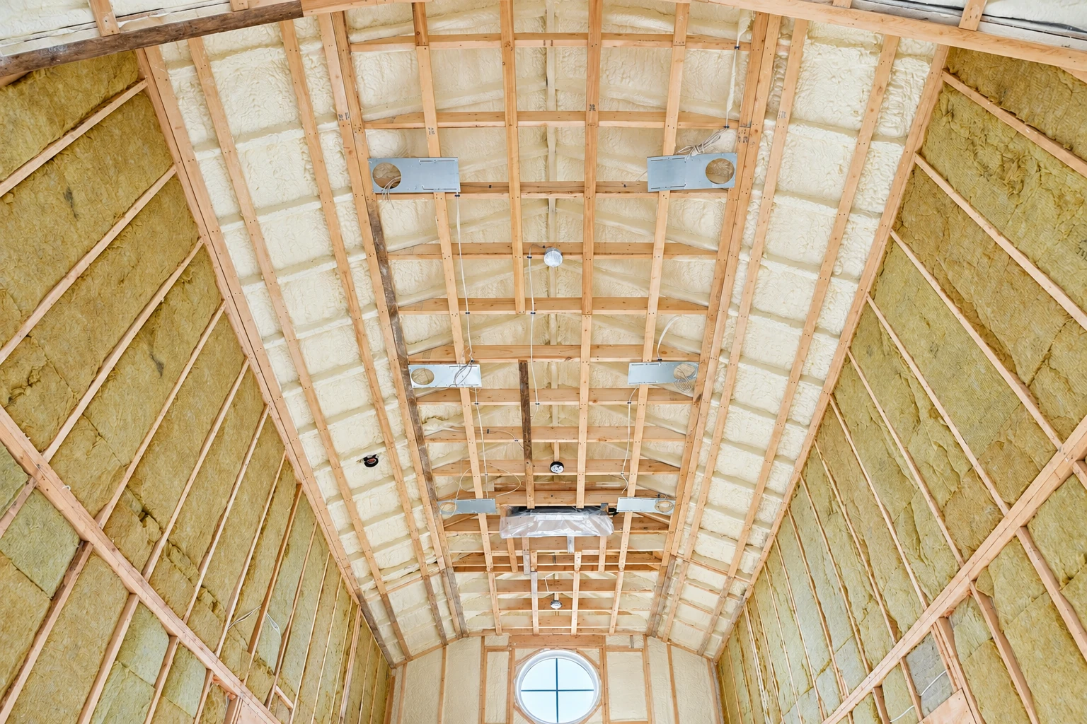 An unfinished vaulted ceiling has pale spray foam between wooden rafters and yellow insulation along the side walls.