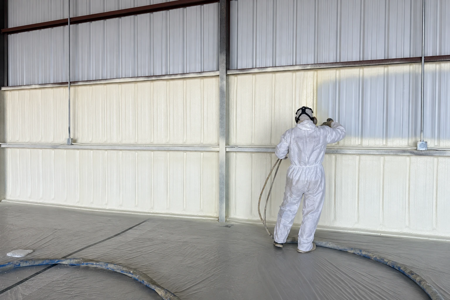 A worker in white protective coveralls applies spray foam insulation to the metal wall of a large commercial building.