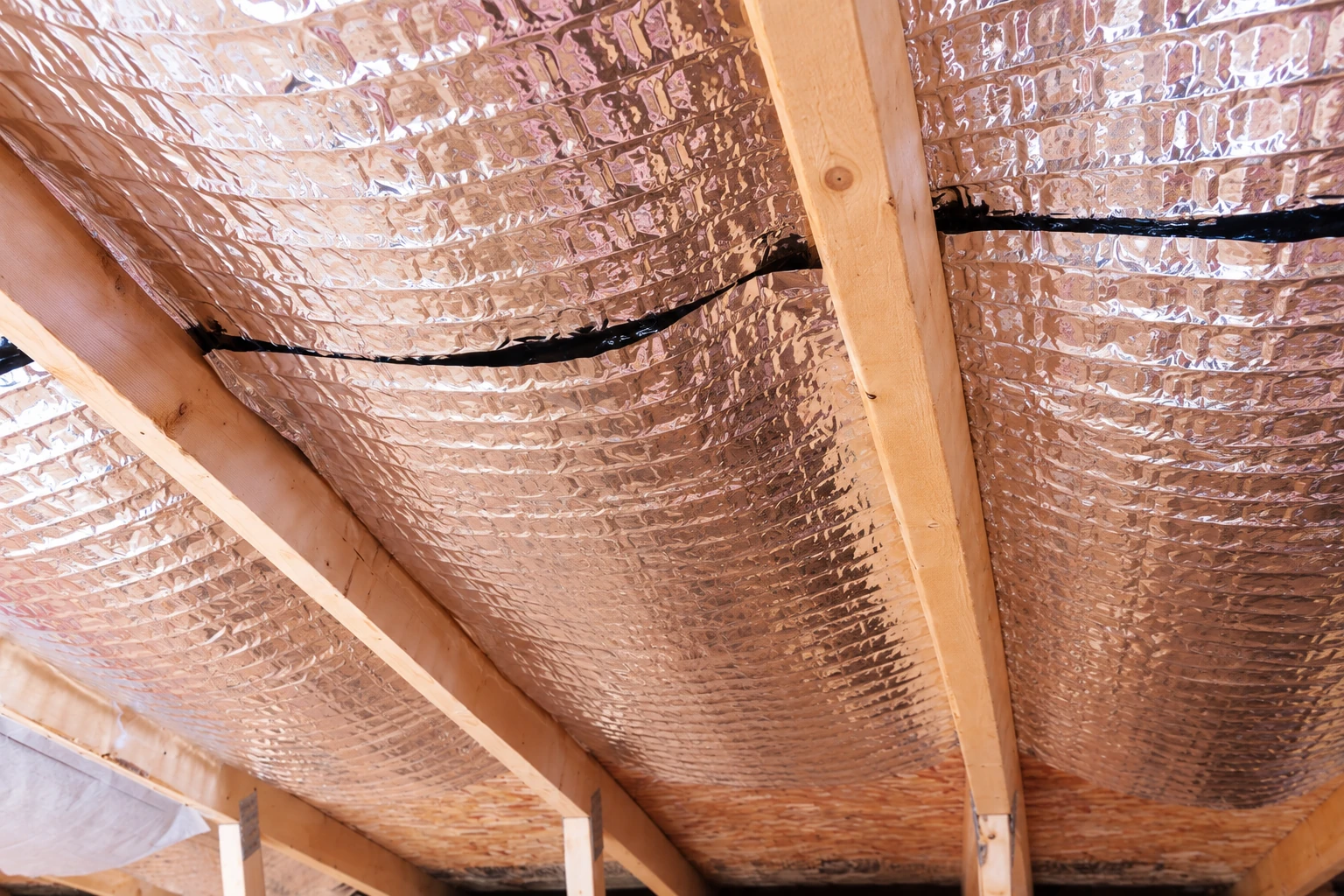 Reflective foil vapor barrier covers the underside of a timber-framed roof, with dark seams between sections.