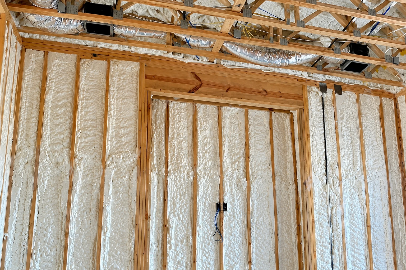 Exposed wall framing filled with white spray foam insulation, with ductwork running above.